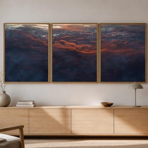 May include: A triptych wall art set featuring a high-altitude aerial view of dense clouds at sunset, with deep blue and dark purple shadows contrasted by vibrant orange and red light illuminating the cloud tops, framed in light wood.