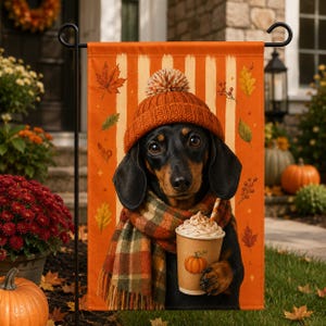 Puede incluir: Una bandera de jardín rectangular con un perro salchicha negro y fuego que lleva un gorro de punto naranja con pompón y una bufanda a cuadros. El perro sostiene un vaso con nata montada y una ilustración de calabaza sobre un fondo de rayas.