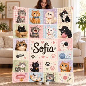 May include: A soft fleece blanket featuring a grid of various cartoon cats, yarn balls, and fish bones. The center panel displays the name Sofia in large black script, surrounded by pink hearts and paw prints on a light cream background.