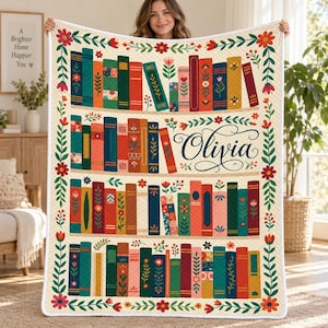 Puede incluir: Una colcha de color crema con un patrón de estantería con lomos de libros coloridos, motivos florales y el nombre Olivia escrito en letra cursiva negra. El borde está decorado con flores y hojas rojas y verdes.