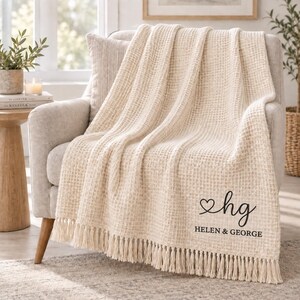 May include: A cream-coloured waffle-knit throw blanket featuring a fringe trim along the bottom edge. The blanket is personalised with a black graphic of a heart and the lowercase letters hg, followed by the text HELEN & GEORGE in capital letters.