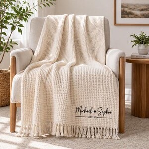May include: A cream-coloured waffle-knit throw blanket with fringe trim on the bottom edge. The blanket features black printed text that reads Michael, a heart symbol, Sophia, and the text EST. 2026 centred below the names.