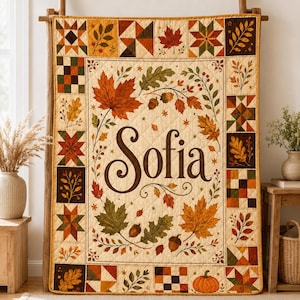 Pode incluir: Uma colcha retangular com um painel central creme com o nome Sofia em letra cursiva castanho-escura, rodeado por folhas de outono, bolotas e uma abóbora. A borda consiste em quadrados de patchwork com padrões de estrelas e folhagens.