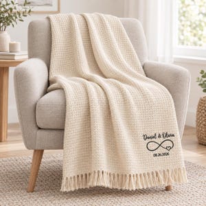 May include: A cream-coloured waffle-knit throw blanket with fringe trim. The blanket features black embroidered text that reads Daniel & Olivia, an infinity symbol with a small heart, and the date 08.26.2026 printed on the lower right corner.