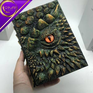 May include: A detailed, green and brown dragon-themed book with an orange eye. The cover features a textured design of scales, spikes, and a dragon's eye. The book is held in a hand.