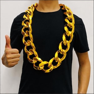 Könnte beinhalten: Eine große, goldfarbene Kette mit übergroßen Gliedern. Die Kette wird um den Hals getragen und hängt über einem schwarzen T-Shirt. Die Person zeigt einen Daumen hoch.