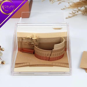 May include: A clear acrylic box displaying a 3D paper sculpture of a Roman coliseum. The sculpture is made of layered paper in shades of brown and beige, with a small bench and tree. The box has a lid and the Snap Design logo.