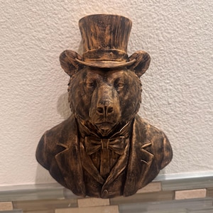 Puede incluir: Busto de oso de color bronce que lleva un sombrero de copa, una pajarita y una chaqueta. La cabeza del oso está detallada con textura de pelaje, y el busto está montado en una pared. La escultura tiene una estética vintage y antigua.
