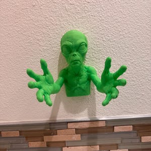 May include: A green alien sculpture with outstretched arms, mounted on a white wall. The alien has a detailed face and textured skin. The sculpture is above a decorative tile border.