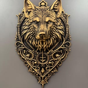 Può includere: Scultura dettagliata della testa di un lupo dorata con dettagli intricati, su sfondo scuro. Il volto del lupo è centrato, circondato da una cornice ornata in stile gotico con dettagli di teschi e croci. La scultura è probabilmente in resina o materiale simile.