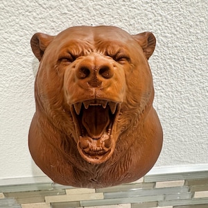 Könnte beinhalten: Eine braune Bärenkopfskulptur mit offenem Maul, die scharfe Zähne zeigt. Das Fell des Bären ist strukturiert, und die Skulptur ist an einer weißen Wand befestigt. Die Augen des Bären sind geschlossen, und es ist ein dekorativer Gegenstand.