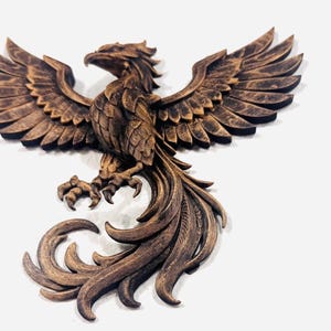 May include: A bronze-colored phoenix sculpture with outstretched wings and detailed feathering. The bird is perched, with its talons visible, and its tail feathers are elegantly curled. The sculpture is mounted on a white background.