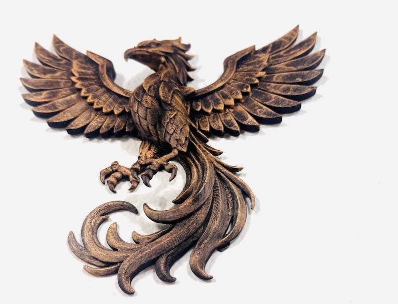 May include: A bronze-colored phoenix sculpture with outstretched wings and detailed feathering. The bird is perched, with its talons visible, and its tail feathers are elegantly curled. The sculpture is mounted on a white background.