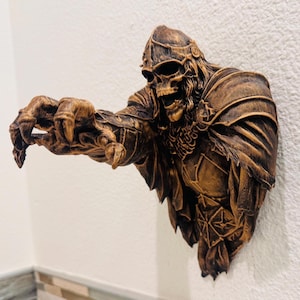 May include: A detailed, brown-toned wall sculpture of a skeletal warrior in ornate armor, with a helmet and outstretched hand. The sculpture is mounted on a textured white wall, showcasing intricate details and a menacing pose.