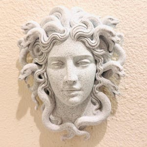 K&ouml;nnte beinhalten: Graue Medusa-Kopf-Skulptur mit Schlangen als Haare, an einer hellbeigen Wand befestigt. Die Skulptur ist ein klassisches Kunstwerk mit geschlossenen Augen und einem ruhigen Ausdruck. Ein dekoratives Element.