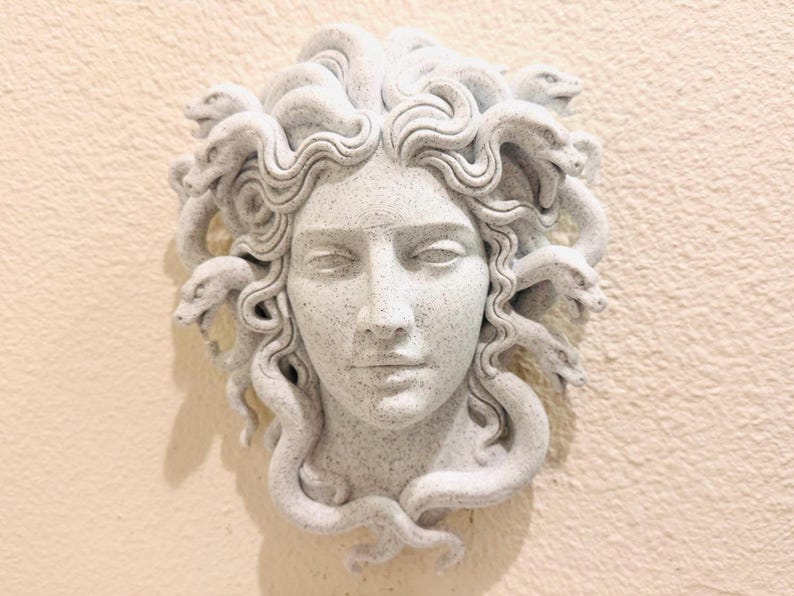 K&ouml;nnte beinhalten: Graue Medusa-Kopf-Skulptur mit Schlangen als Haare, an einer hellbeigen Wand befestigt. Die Skulptur ist ein klassisches Kunstwerk mit geschlossenen Augen und einem ruhigen Ausdruck. Ein dekoratives Element.
