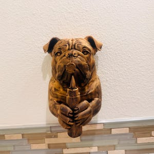 May include: A detailed, brown bulldog sculpture holding a wooden object. The bulldog's face is intricately carved, with visible wrinkles and a serious expression. The sculpture is mounted on a wall, showcasing its detailed craftsmanship.