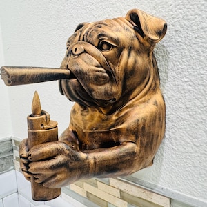 May include: A wall-mounted sculpture of a bulldog with a cigar in its mouth and a lighter in its paws. The sculpture is finished in shades of brown and gold, with detailed textures. The bulldog's expression is stoic.