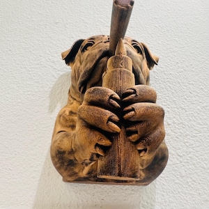 May include: A brown sculpture of a bulldog holding a long, cylindrical object to its mouth. The bulldog's paws are wrapped around the object. The sculpture is mounted on a white wall.