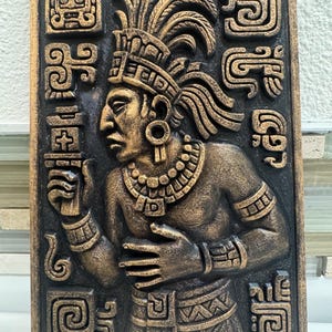 May include: A rectangular wall plaque featuring a relief of a Mayan figure in profile, adorned with a headdress and jewelry. The figure is rendered in bronze tones against a dark background, surrounded by Mayan glyphs. The plaque has a textured surface.