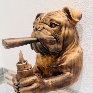 May include: A bronze-colored bulldog sculpture with a cigar in its mouth and a lighter in its paw. The bulldog has detailed wrinkles and is mounted on a white wall. The sculpture is a decorative item.