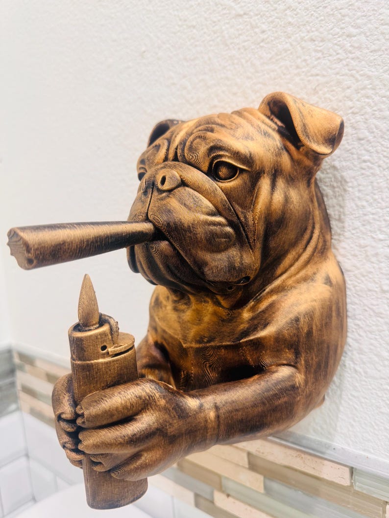 May include: A bronze-colored bulldog sculpture with a cigar in its mouth and a lighter in its paw. The bulldog has detailed wrinkles and is mounted on a white wall. The sculpture is a decorative item.