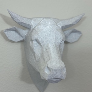 Peut inclure: Sculpture géométrique d'une tête de vache grise claire, avec des cornes et des oreilles pointues. La sculpture est fixée au mur, mettant en valeur un design moderne et minimaliste. La tête de vache est faite d'un matériau texturé.