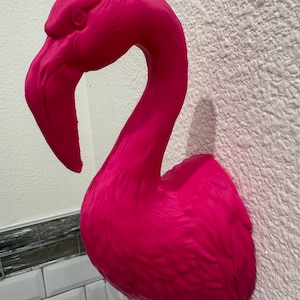 Könnte beinhalten: Eine leuchtend pinkfarbene Flamingo-Wandskulptur. Der Flamingo hat einen langen, gebogenen Hals und einen detaillierten Schnabel. Die Skulptur ist an einer strukturierten weißen Wand befestigt und verleiht jedem Raum eine tropische Note.