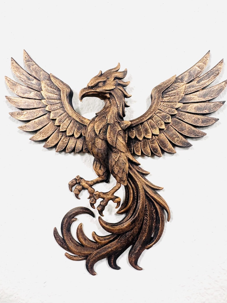 May include: A bronze-colored phoenix sculpture with outstretched wings and detailed feathering. The bird's talons are visible, and the tail feathers curl elegantly. This wall art piece is a decorative item.