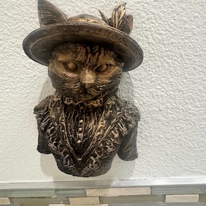May include: A bronze-toned cat sculpture, featuring a hat with a feather and a detailed ruffled collar. The cat's face is intricately sculpted. The bust is mounted on a white wall, offering a vintage aesthetic. The hat and collar add a touch of elegance.