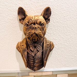 May include: A bronze-colored wall sculpture of a French Bulldog wearing a suit jacket and bow tie. The dog's face is detailed with wrinkles and expressive eyes. The sculpture is mounted on a white wall.