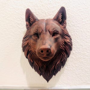 Könnte beinhalten: Detaillierte, braune Wolfskopfskulptur an einer weißen Wand. Die Skulptur weist komplizierte Details des Fells, der Augen und der Nase des Wolfs auf, wodurch ein realistisches und strukturiertes Aussehen entsteht. Ein dekoratives Kunstwerk für die Inneneinrichtung.