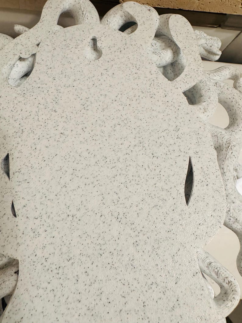 K&ouml;nnte beinhalten: Ein wei&szlig;es, gesprenkeltes Dekorationsobjekt mit einem Medusenhaupt-Design. Die Skulptur weist komplizierte Details auf, darunter sich windende Schlangen und eine strukturierte Oberfl&auml;che. Das Objekt hat ein gesprenkeltes Muster aus schwarzen und grauen Punkten.