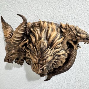 May include: A detailed, three-headed mythical creature sculpture with horns and a textured, layered design. The sculpture is primarily bronze and brown, mounted on a white wall. The creature's heads are intricately carved, showcasing a blend of animal and fantastical features.