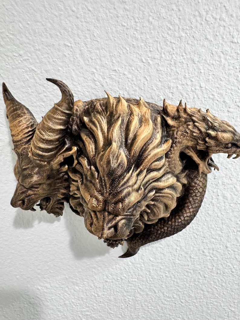 May include: A detailed, three-headed mythical creature sculpture with horns and a textured, layered design. The sculpture is primarily bronze and brown, mounted on a white wall. The creature's heads are intricately carved, showcasing a blend of animal and fantastical features.
