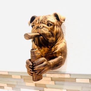 May include: A bronze-colored bulldog wall sculpture with a cigar in its mouth and a lighter in its paw. The sculpture is mounted on a white wall with a decorative tile border. The bulldog's features are detailed, and the overall effect is rustic.