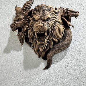 May include: A bronze-colored wall sculpture depicting a three-headed mythical creature. The heads include a lion, a goat, and a dragon, each with open mouths and detailed textures. The sculpture is mounted on a white wall.