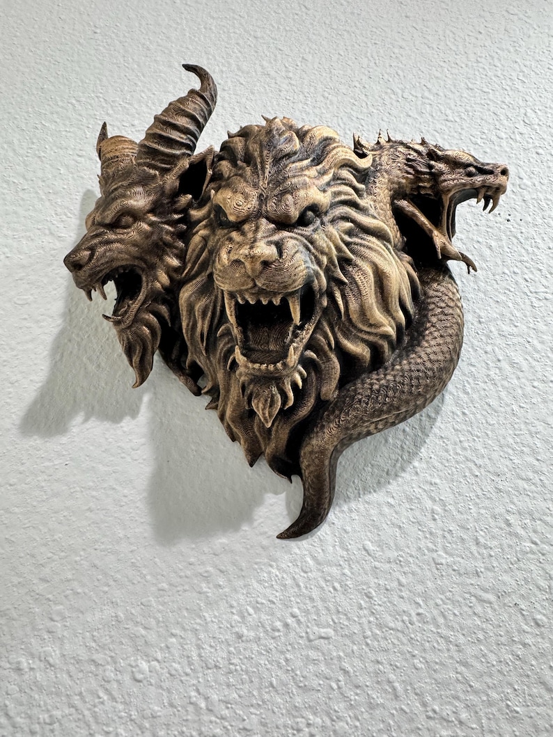 May include: A bronze-colored wall sculpture depicting a three-headed mythical creature. The heads include a lion, a goat, and a dragon, each with open mouths and detailed textures. The sculpture is mounted on a white wall.
