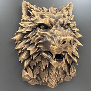 May include: A bronze-coloured wolf head sculpture with a textured, spiky mane and open mouth revealing teeth. The sculpture has a fierce expression and is mounted on a grey surface. The artwork is a decorative piece.