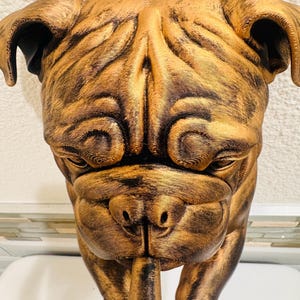 May include: A bronze-colored sculpture of a bulldog's head, with detailed wrinkles and folds. The dog's face is centered, with a pipe extending from its mouth. The sculpture has a metallic sheen, suggesting a painted or coated finish. The bulldog is a popular breed.