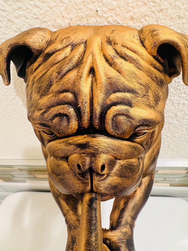 May include: A bronze-colored sculpture of a bulldog's head, with detailed wrinkles and folds. The dog's face is centered, with a pipe extending from its mouth. The sculpture has a metallic sheen, suggesting a painted or coated finish. The bulldog is a popular breed.