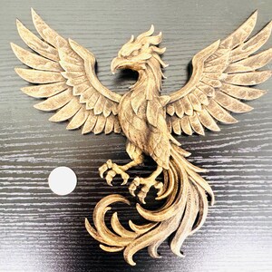 May include: A detailed, golden-brown phoenix sculpture with outstretched wings and a flowing tail. The bird is intricately carved, showcasing feathers and a fierce expression. The sculpture is placed on a dark, wood-grain surface.