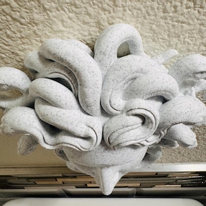 K&ouml;nnte beinhalten: Eine abstrakte wei&szlig;e Skulptur mit einem gesprenkelten Muster. Die Skulptur hat eine komplexe, organische Form mit abgerundeten Formen und einer spitzen Basis. Das St&uuml;ck scheint aus einem festen Material zu bestehen und ist vor einem neutralen Hintergrund platziert.