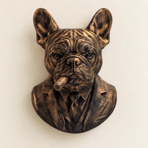 May include: A bronze-toned wall sculpture depicting a French Bulldog wearing a suit, tie, and collared shirt. The dog has a cigar in its mouth and features a textured, weathered finish with dark highlights across its face and clothing.