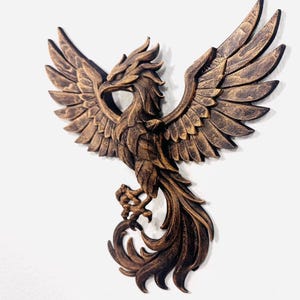 May include: A detailed, bronze-colored phoenix sculpture with outstretched wings. The bird's feathers and talons are intricately carved, and the sculpture is mounted on a white background. The phoenix is a symbol of rebirth and renewal.