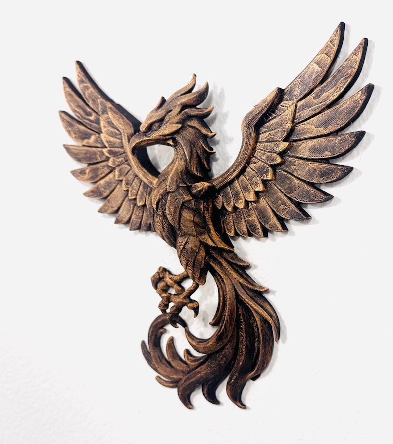 May include: A detailed, bronze-colored phoenix sculpture with outstretched wings. The bird's feathers and talons are intricately carved, and the sculpture is mounted on a white background. The phoenix is a symbol of rebirth and renewal.
