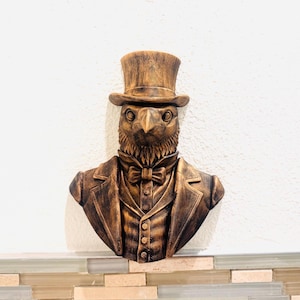 May include: A bronze-colored wall sculpture of an anthropomorphic bird wearing a top hat, bow tie, and suit jacket. The sculpture has detailed textures and a vintage aesthetic, suitable for home decor.