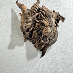 May include: A detailed, three-headed mythical creature wall sculpture. The sculpture features a goat-like head with horns, a lion-like head, and a serpent-like head. The sculpture is a brown color and is mounted on a white wall.