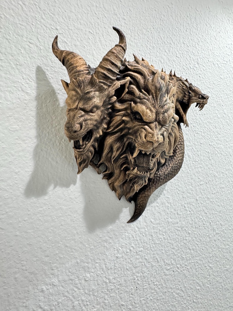 May include: A detailed, three-headed mythical creature wall sculpture. The sculpture features a goat-like head with horns, a lion-like head, and a serpent-like head. The sculpture is a brown color and is mounted on a white wall.