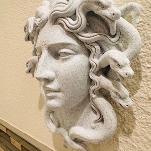 K&ouml;nnte beinhalten: Eine detaillierte, gesprenkelte, graue Skulptur des Medusenhauptes, mit aufwendig geschnitzten Schlangen anstelle von Haaren. Die Skulptur ist an einer hellen Wand befestigt und zeigt das klassische Kunstwerk.